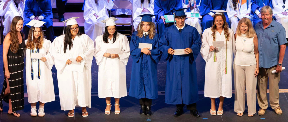 A group of graduates and faculty standing on a stage during a graduation ceremony, wearing white and blue caps and gowns. Students are seated in the background on steps.